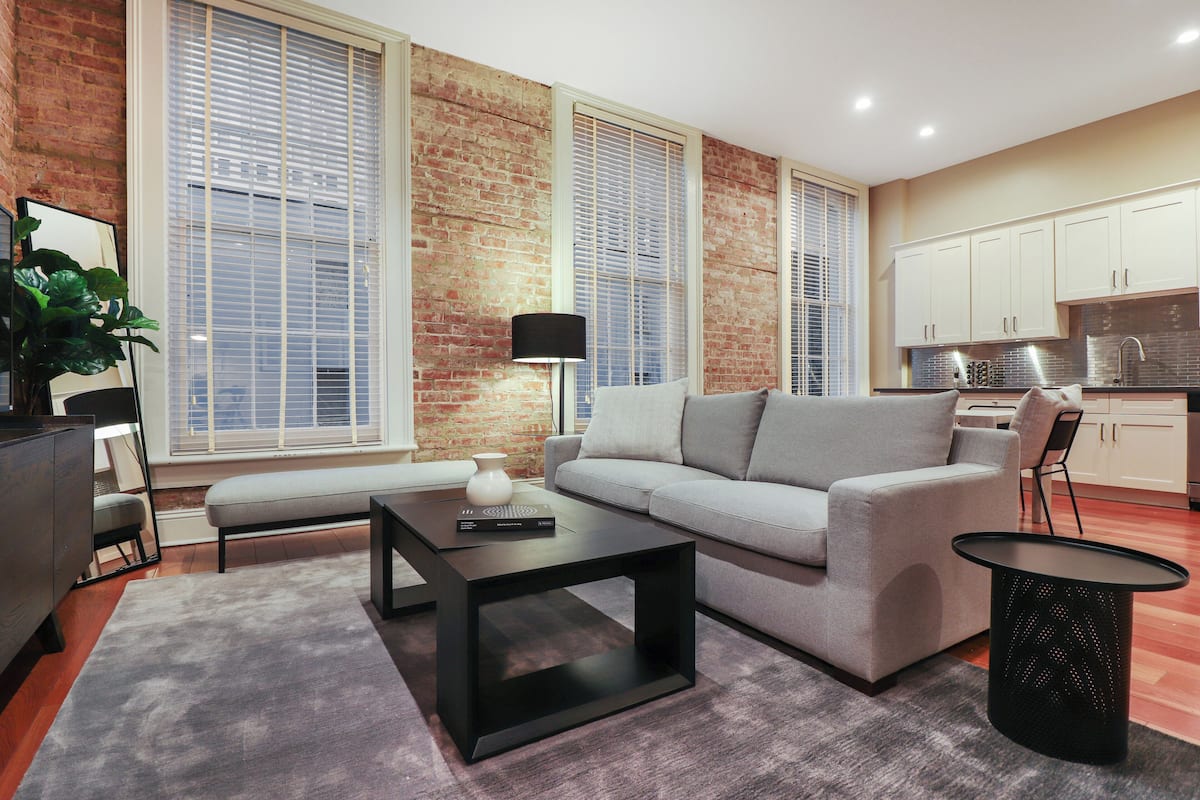 Uma sala de estar moderna com um sofá cinzento, uma mesa de centro preta e uma pequena mesa lateral. Apresenta uma parede de tijolos e janelas amplas com persianas.
