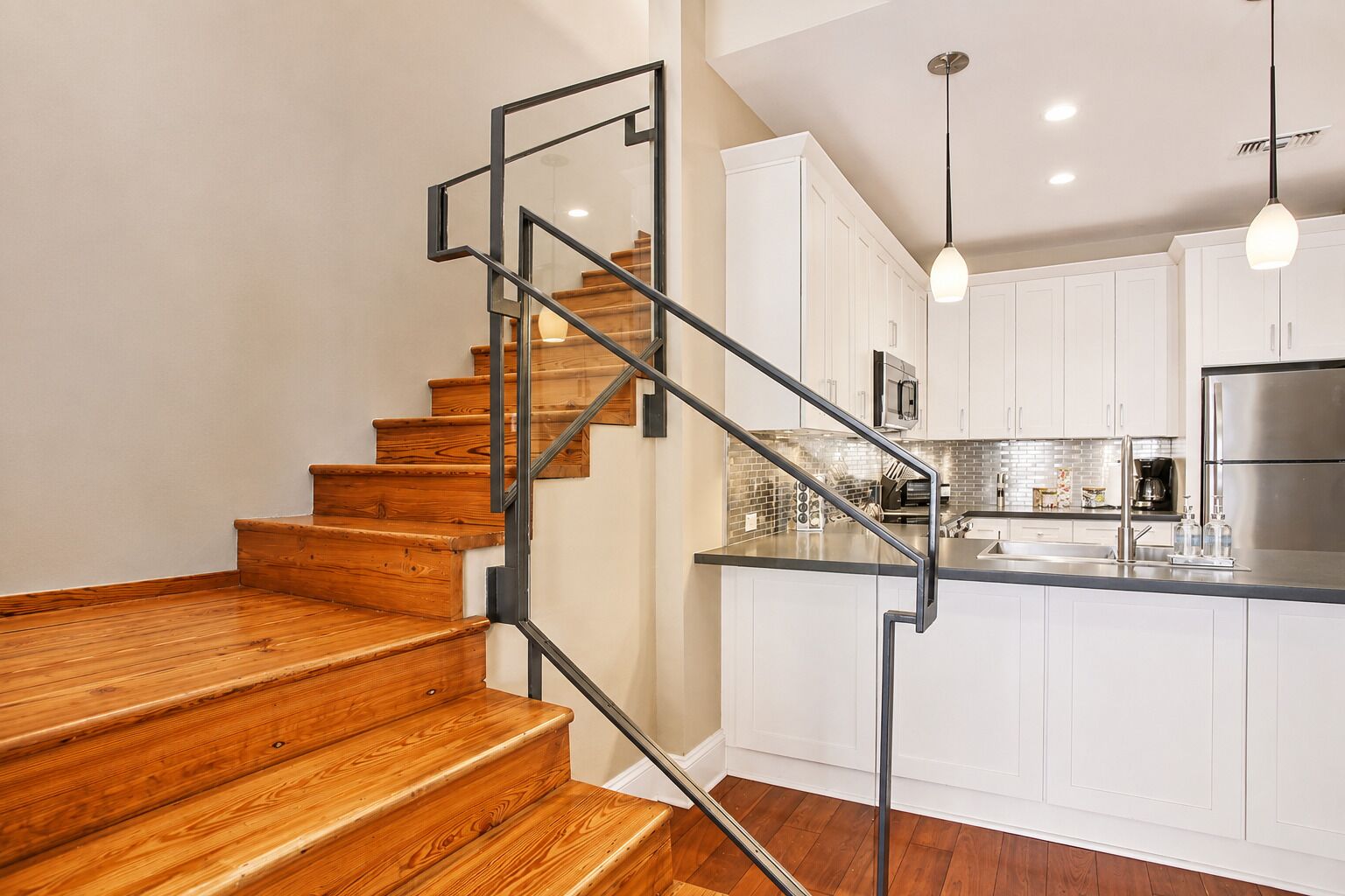A modern kitchen with white cabinets, a stainless steel refrigerator, and a wooden staircase with metal railings.