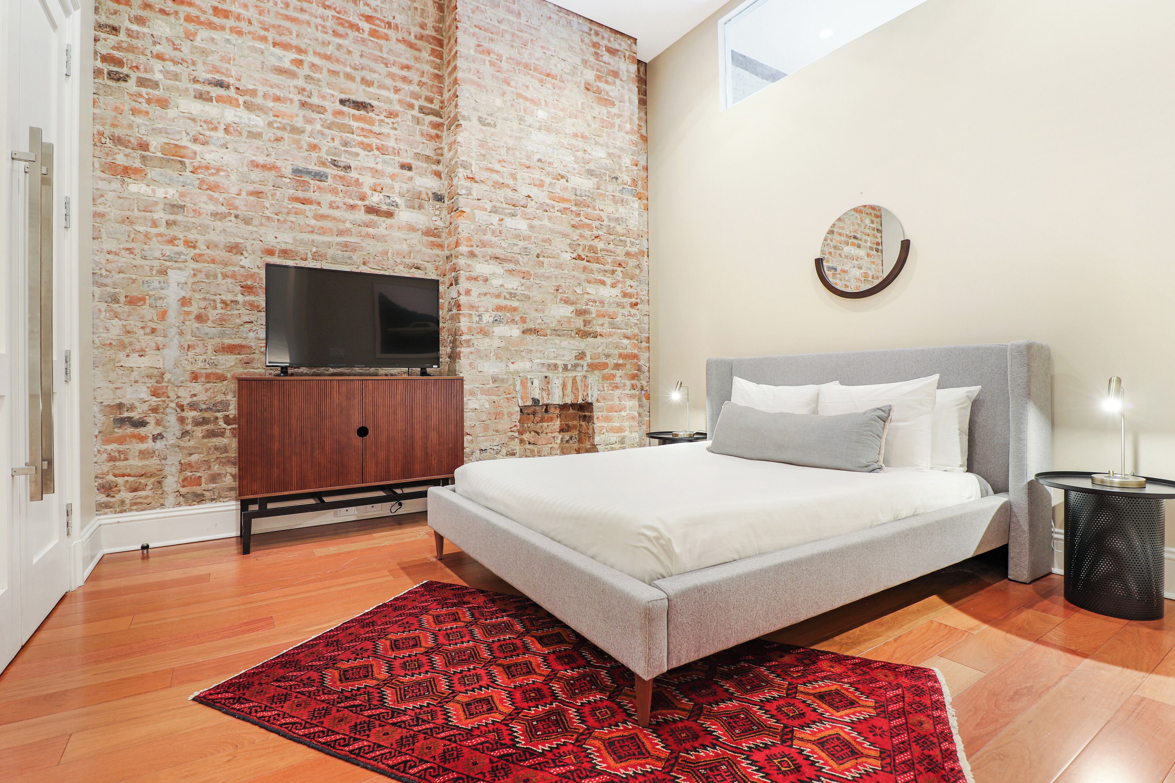 A bedroom with a brick wall, a bed, a TV on a wooden cabinet, and a red patterned rug.