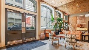 A spacious lobby with a leather sofa, patterned chairs, a large potted plant, and a view of the street through large windows. - Roami at Factors Row (New Orleans)