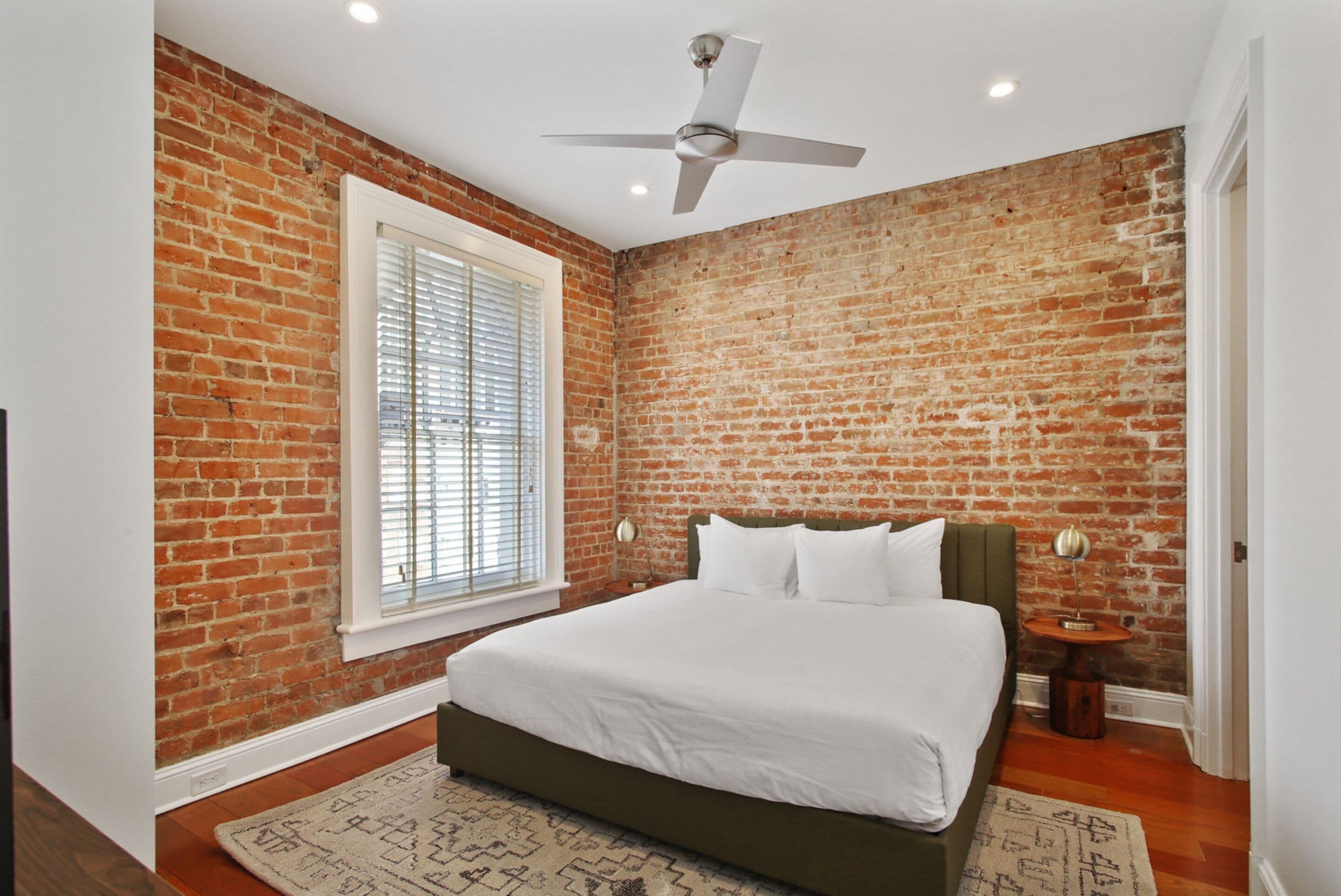 Une chambre avec un mur en briques, un lit, une fenêtre avec des stores, un ventilateur de plafond et une table de chevet avec une lampe.
