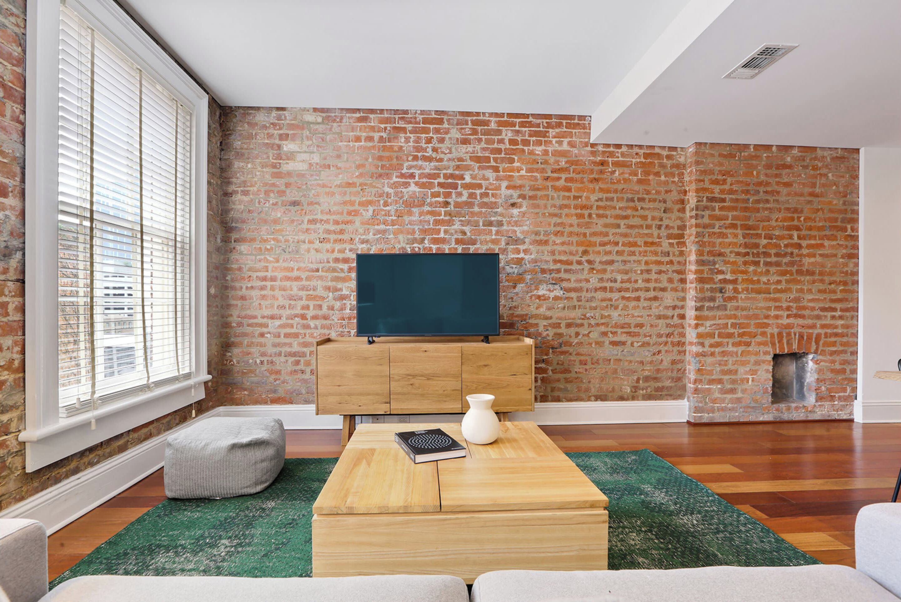 A living room with a brick wall, a flat-screen TV on a wooden cabinet, a green rug, and a white vase.