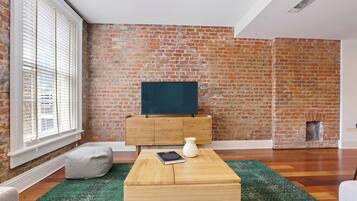 A living room with a brick wall, a flat-screen TV on a wooden cabinet, a green rug, and a white vase.