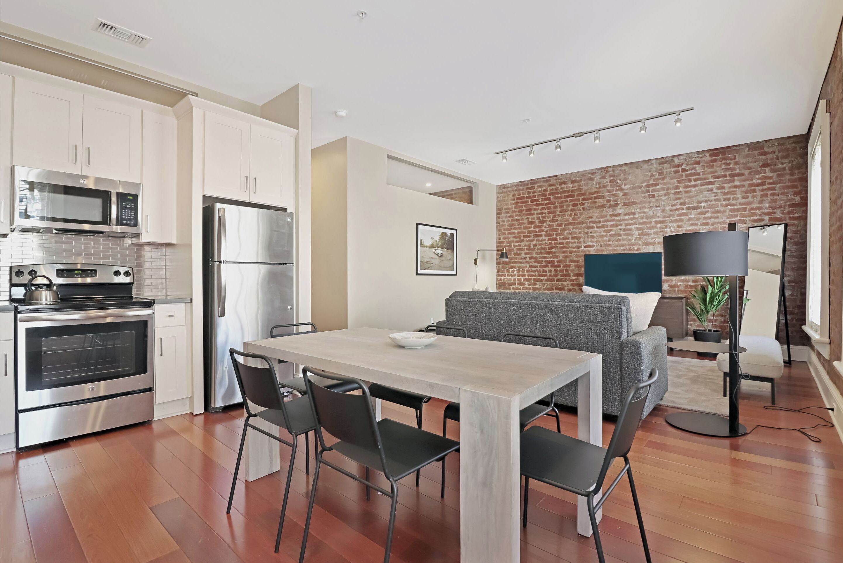 A modern kitchen with stainless steel appliances, a dining area with a light-colored table and chairs, and a living area with a gray sofa and brick wall backdrop.