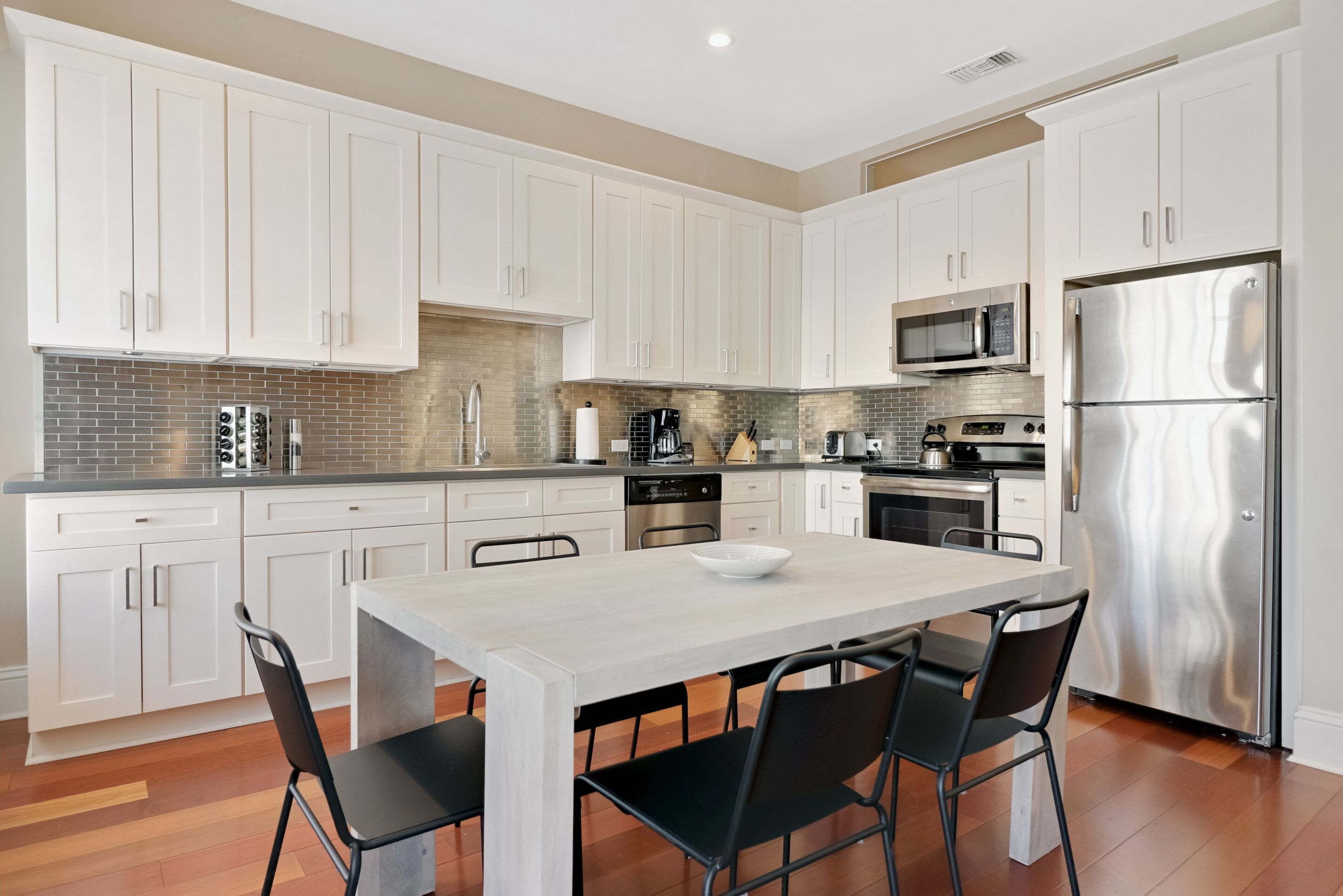 A modern kitchen with white cabinets, stainless steel appliances, and a light-colored dining table with four chairs.