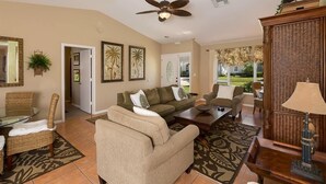 A spacious living room with a sofa, armchairs, a coffee table, and a ceiling fan. There is a dining area with a glass table and chairs, and a decorative wall with framed artwork. - 1650 Galleon Court (Marco Island)