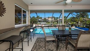 A poolside area with a table and chairs, a ceiling fan, and a view of a pool and palm trees. - 1650 Galleon Court (Marco Island)