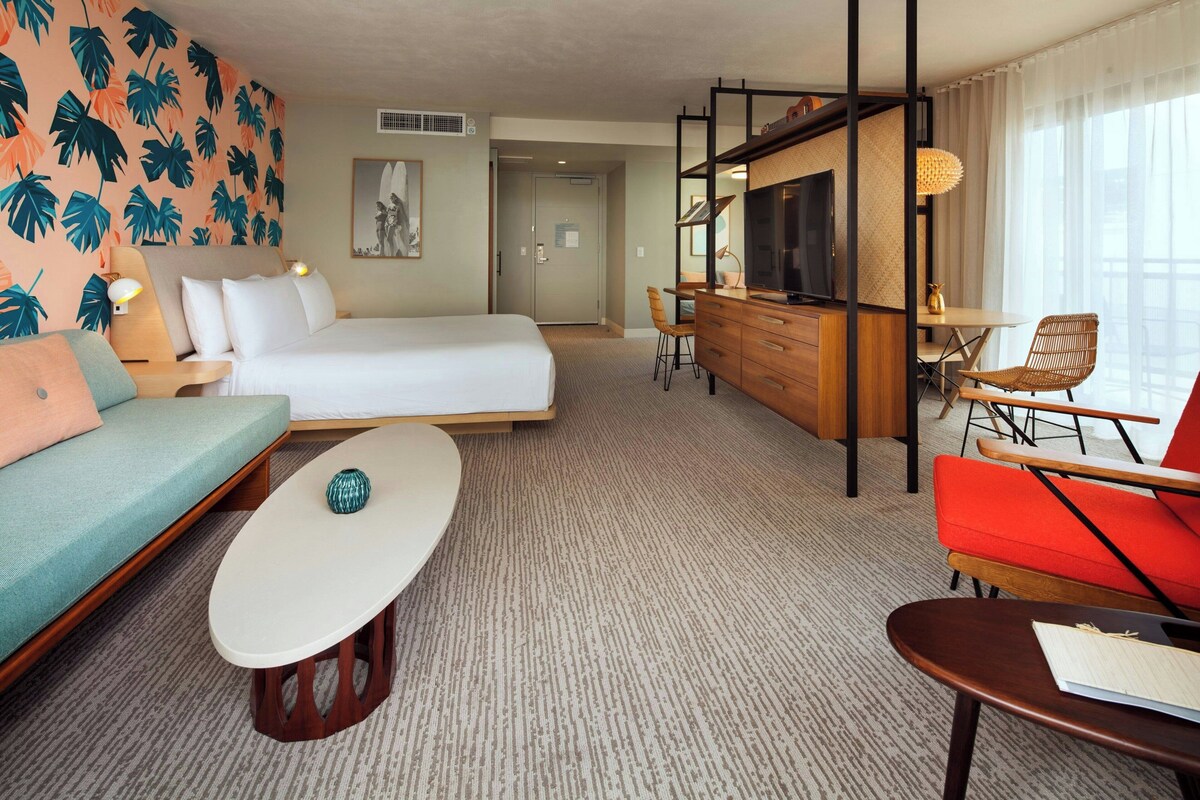 A hotel room with a bed, sofa, armchair, TV, desk, and a large window.