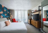 A modern hotel room with a large bed, a TV, a wooden dresser, and a red chair.