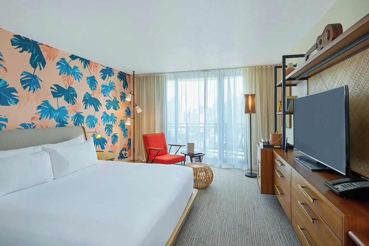 A hotel room with a bed, a television, a desk with a lamp, a chair, and a large window with curtains.