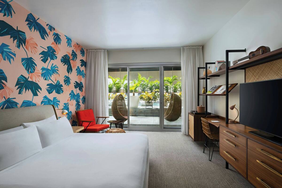 A hotel room with a bed, a desk, a TV, and a view of the outdoors through a sliding glass door.