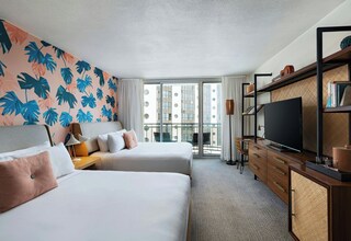 A hotel room with two beds, a TV, a bookshelf, and a balcony with a view of buildings.