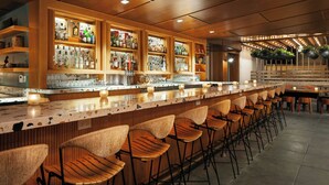 A bar area with a long counter, multiple shelves of bottles, and several wooden bar stools. - The Laylow Waikiki, Autograph Collection (Honolulu)