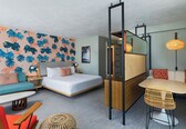 A hotel room with a bed, a sofa, a dining table, and a chair. There is a mirror and a lamp on the wall.