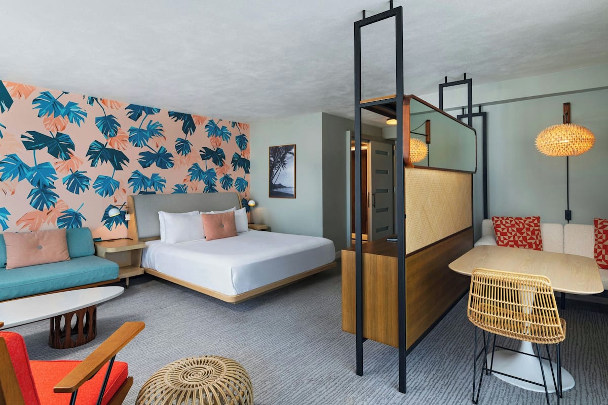A hotel room with a bed, a sofa, a dining table, and a chair. There is a mirror and a lamp on the wall.