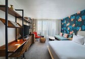 A hotel room with a bed, a desk with a chair, a sofa, a television, and a wall with a tropical leaf pattern.