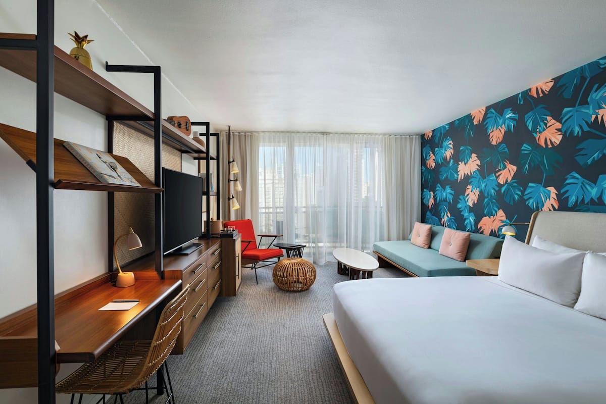 A hotel room with a bed, a desk with a chair, a sofa, a television, and a wall with a tropical leaf pattern.