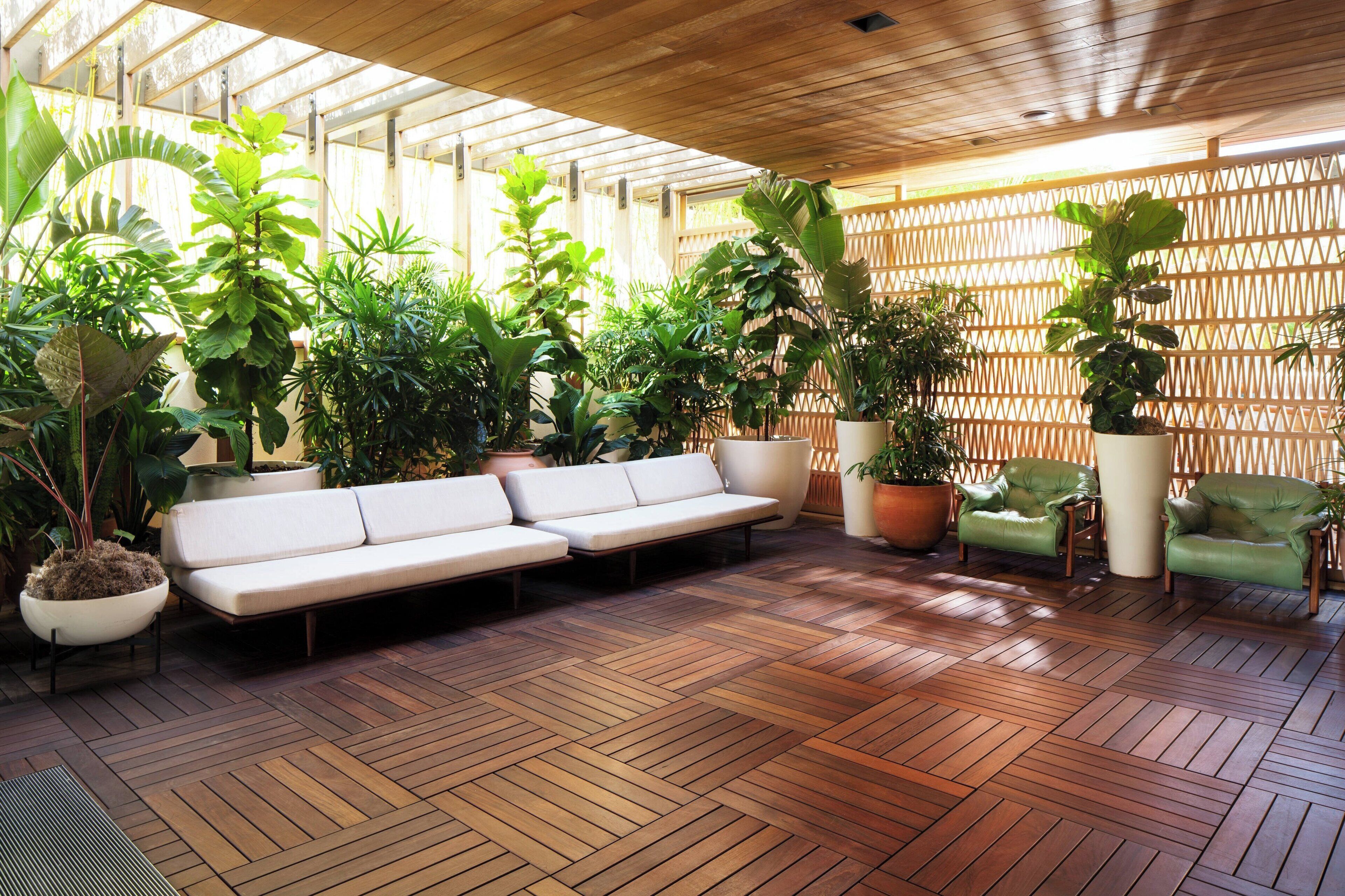 Un espacio al aire libre techado con piso de madera, sofá blanco y sillones verdes, rodeado de plantas en macetas.