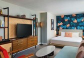 A modern hotel room with a flat-screen TV, wooden furniture, a bed with a tropical leaf wallpaper, and a small table.