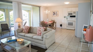 A living room with a sofa, coffee table, and dining area with chairs and a table. There is a kitchen with appliances and a refrigerator.