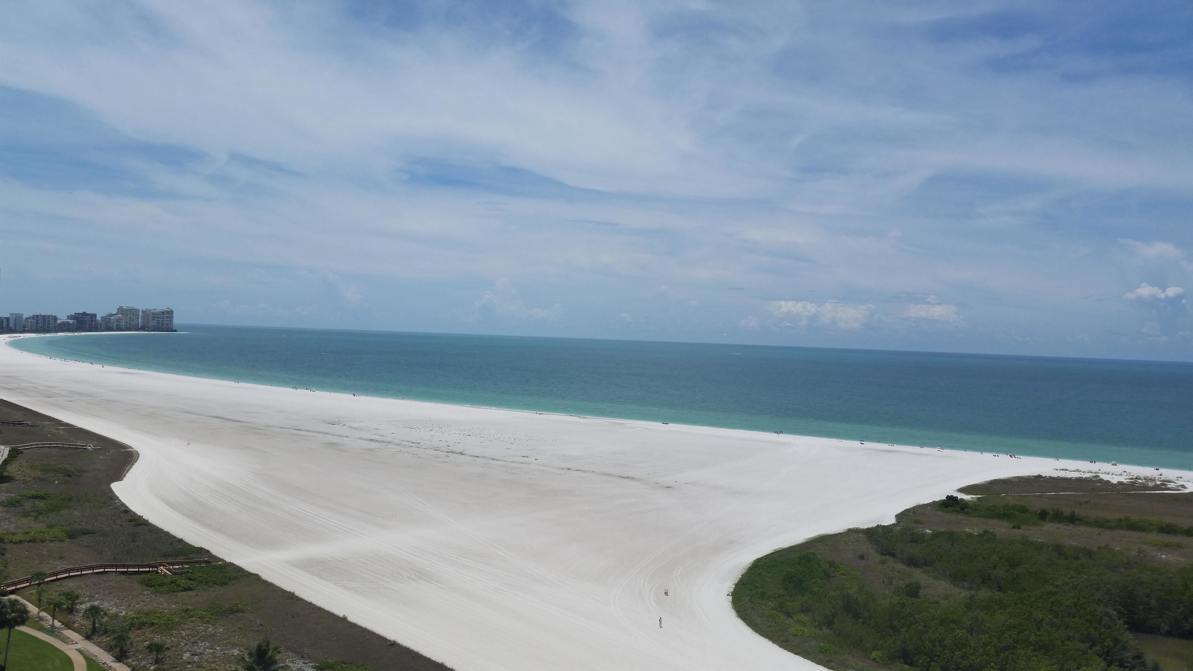 A wide beach with a gentle curve, a clear blue sky, and distant buildings.