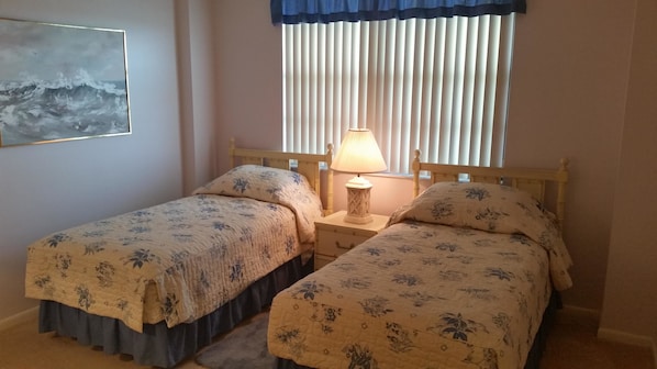 A bedroom with two single beds, a bedside table with a lamp, a window with vertical blinds, and a painting on the wall.