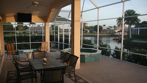 A covered outdoor patio with a dining table and chairs, a pool, and a view of a body of water and buildings in the background. - 431 Edgewater Ct (Marco Island)