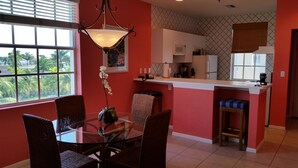 A dining area with a glass table and wicker chairs, a kitchen with white cabinets and a microwave, and a view of palm trees through the window. - Olde Marco Inn 401 (Marco Island)