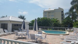 A poolside area with lounge chairs and umbrellas, a pool, and a building in the background. - Grandview 310 (Marco Island)
