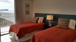 Ein Hotelzimmer mit zwei Betten, einem Nachttisch mit Lampe und Blick auf den Strand.