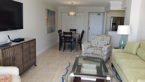 A living room with a TV, a glass coffee table, a sofa, a patterned armchair, a dining area with chairs and a table, and a kitchen area with cabinets and a refrigerator.