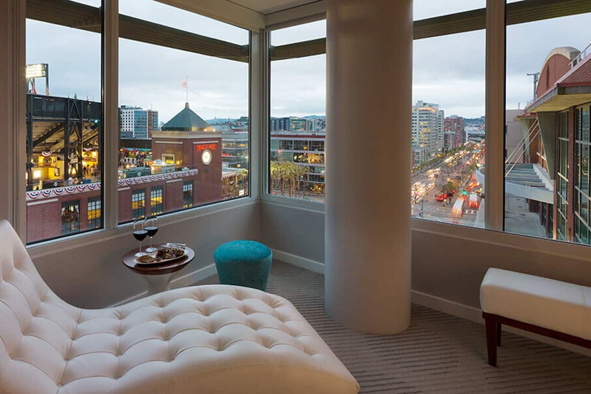 A spacious balcony with a white cushioned chair, a small round table with a tray of food and drinks, and a blue ottoman.