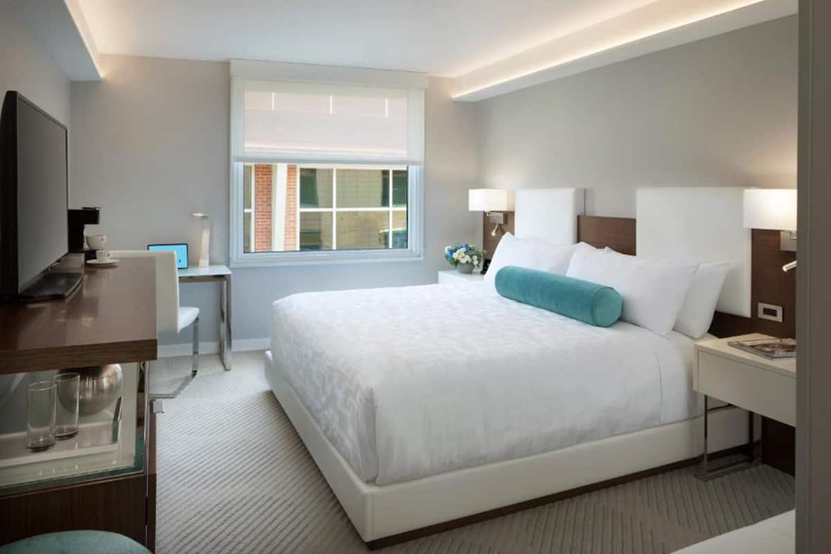 A modern hotel room with a large bed, bedside lamps, a desk with a computer, and a window with blinds.