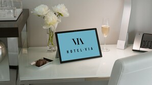 A hotel reception desk with a sign reading "HOTEL VIA", a vase with white flowers, a glass of champagne, and a small plate with a chocolate dessert. - Hotel VIA (San Francisco)