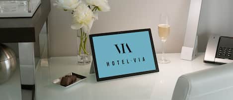 A hotel reception desk with a sign reading "HOTEL VIA", a vase with white flowers, a glass of champagne, and a small plate with a chocolate dessert.