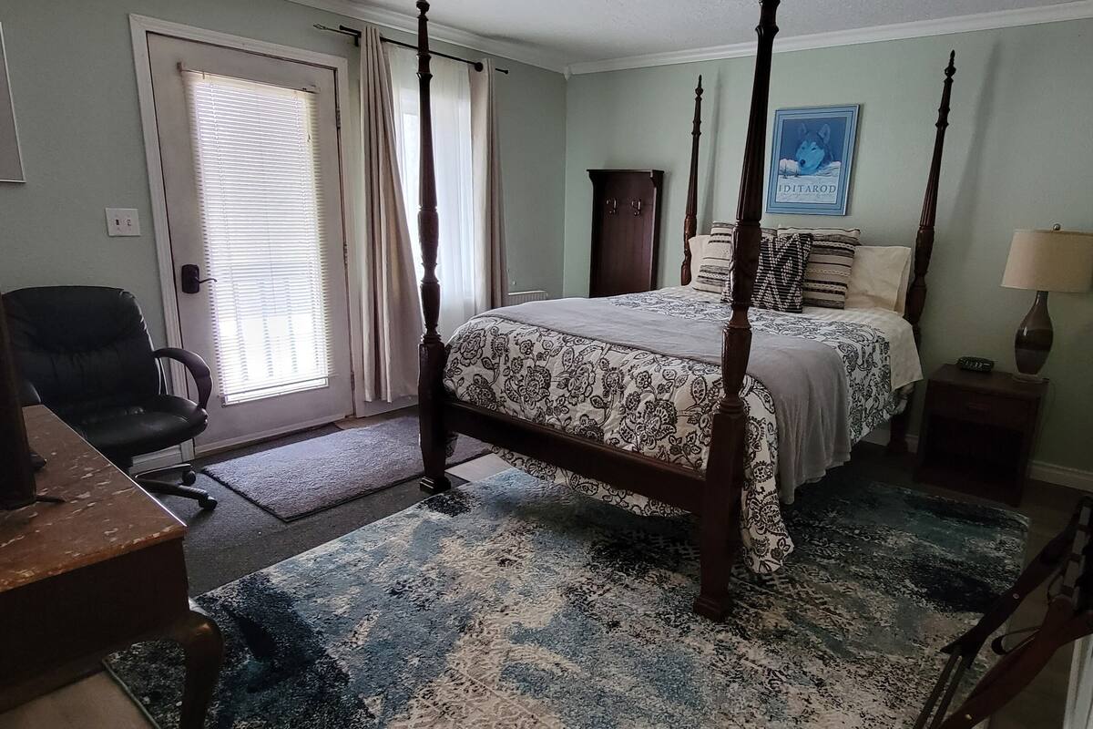 Una cama con dosel con colcha estampada, un escritorio con silla, una mesita de noche con lámpara y un cuadro en la pared.