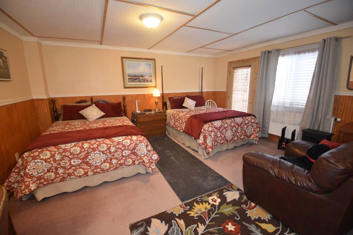 Una habitación doble con dos camas, un cabecero de madera, una mesita de noche, una lámpara, un sillón de cuero y una ventana con cortinas.