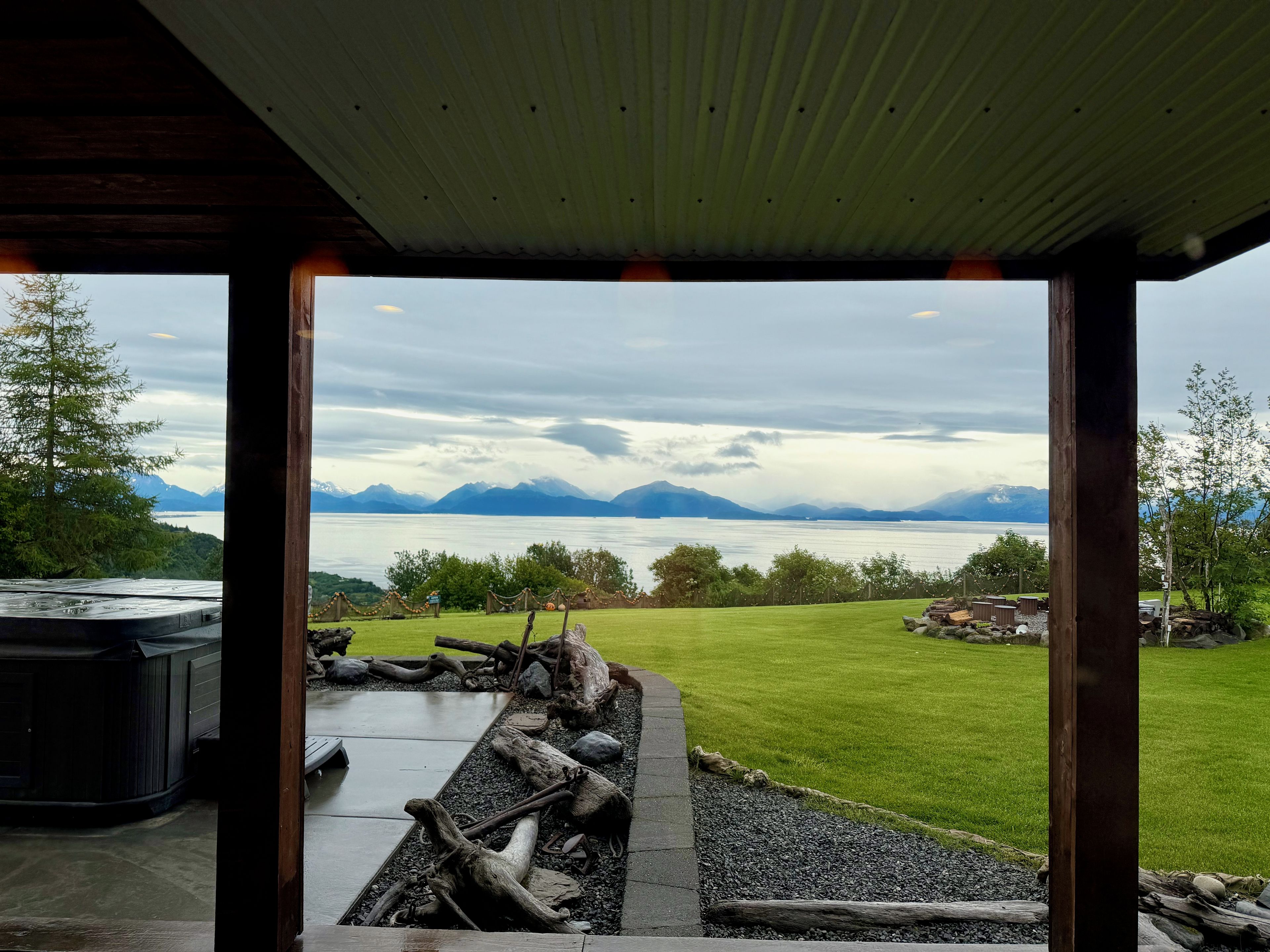 A view from a covered outdoor area with a patio, a hot tub, and a grassy field leading to a body of water with mountains in the distance.