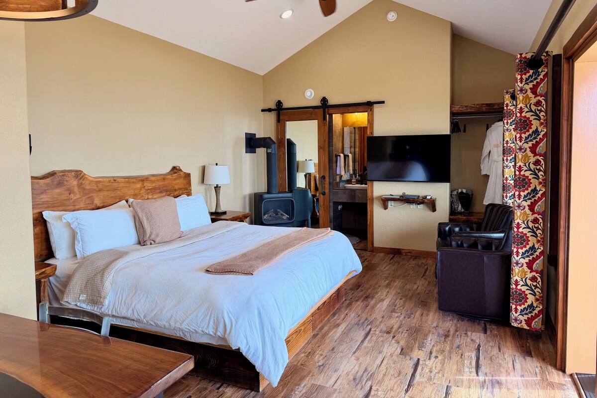 Une chambre à coucher avec un lit, un ventilateur de plafond, une télévision, une cheminée et une salle de bain visible à travers une porte ouverte.