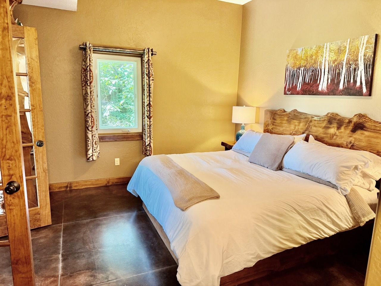 A bedroom with a bed, a window with curtains, a painting on the wall, and a wooden headboard.