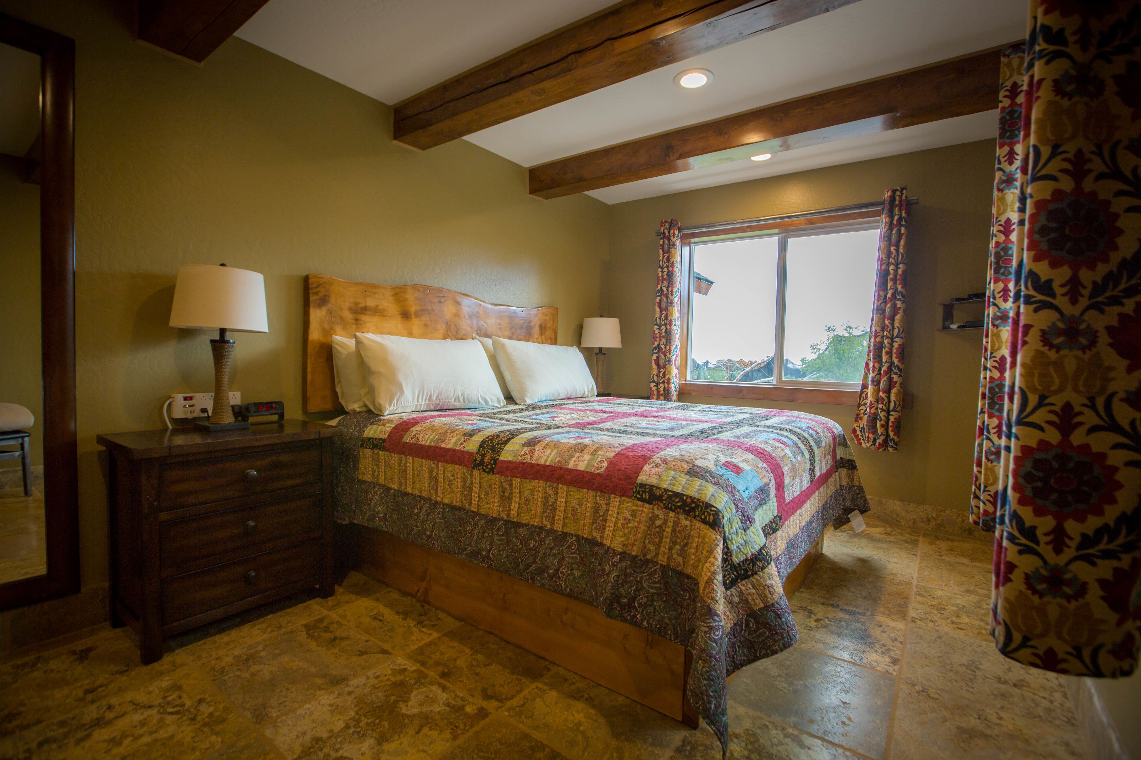A bedroom with a wooden headboard, a large bed with a colorful quilt, two bedside lamps, a dresser, and a window with curtains.