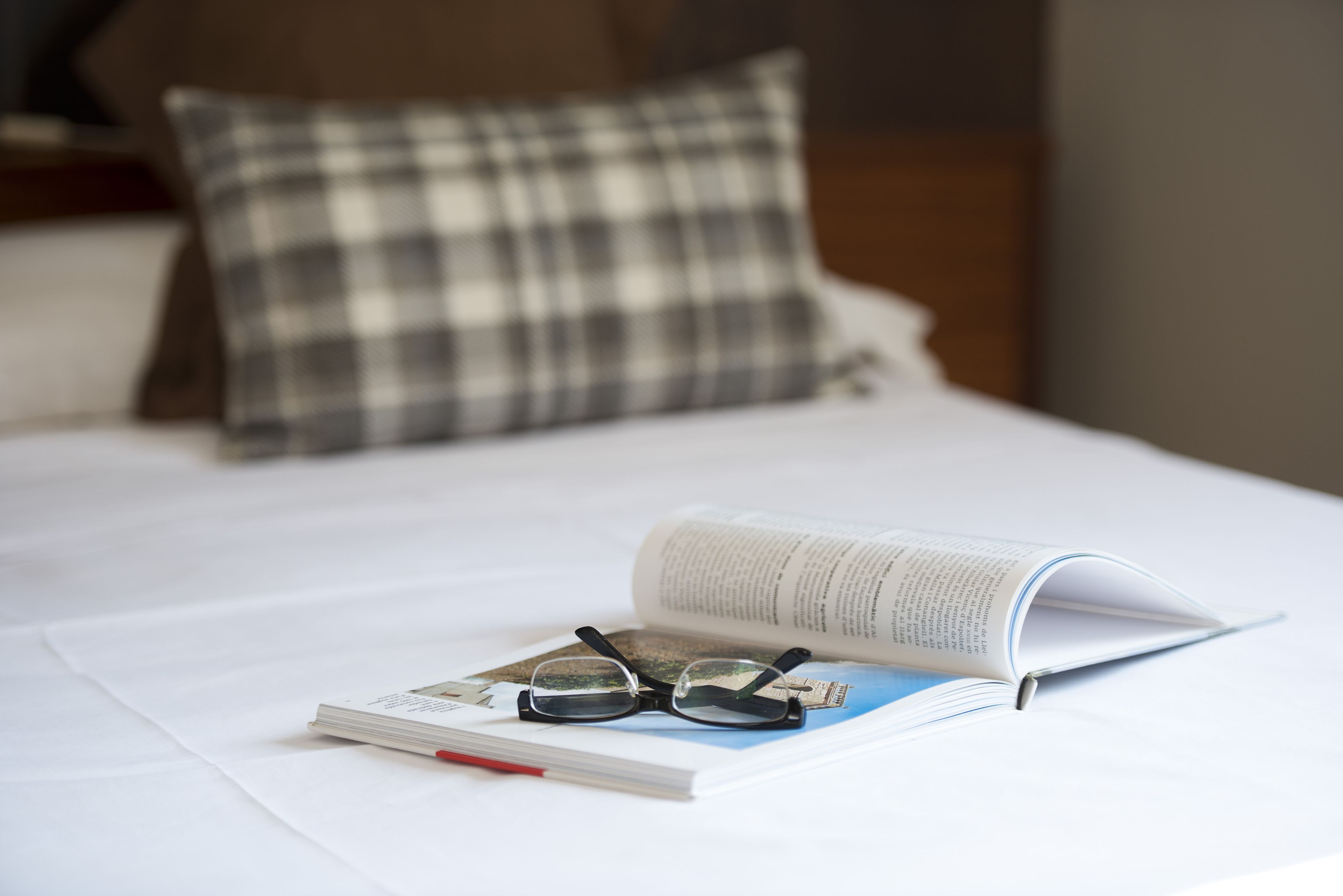 Una cama con un cojín de cuadros, un libro y un par de gafas sobre una sábana blanca.