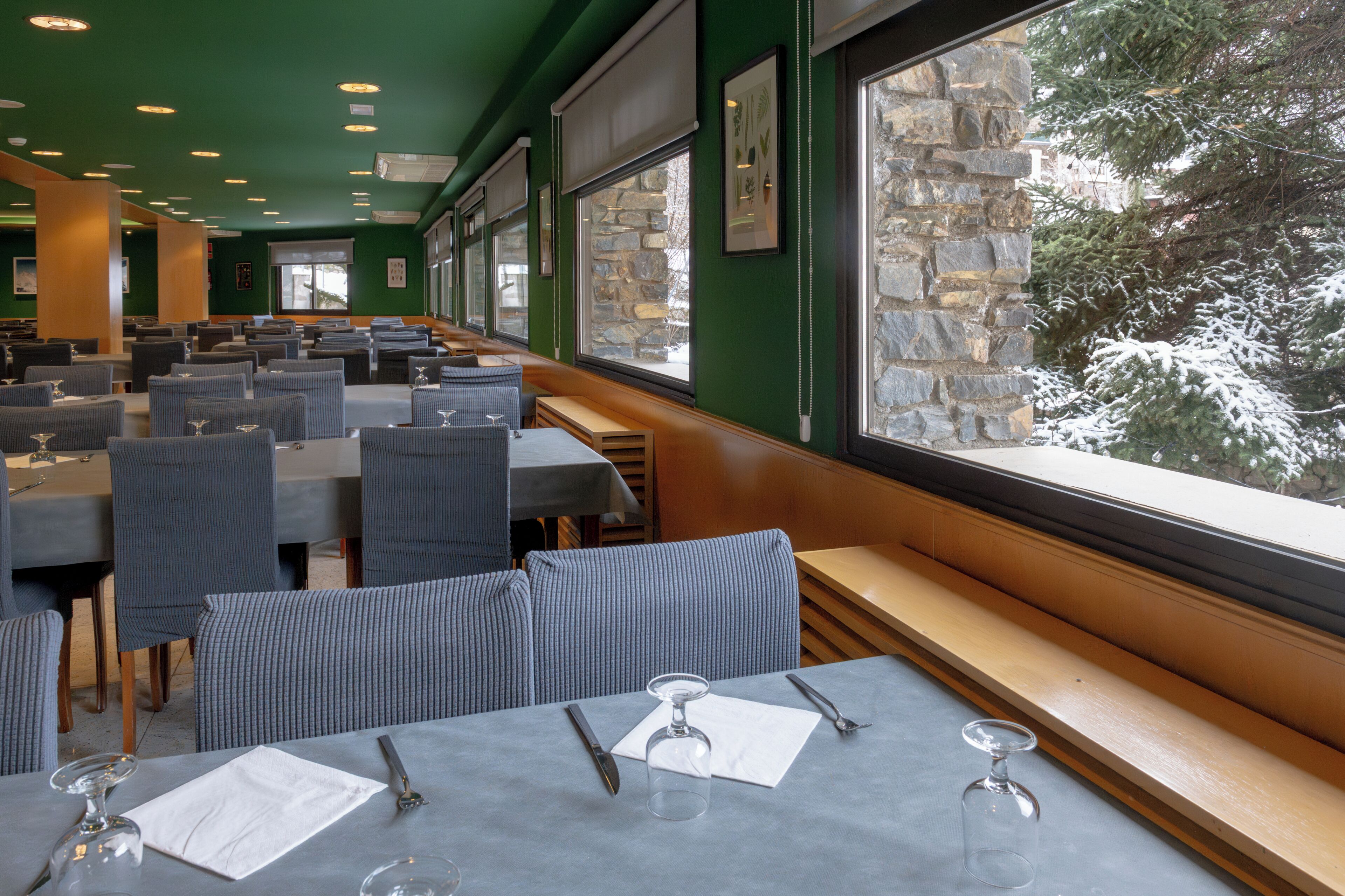 A restaurant with multiple tables and chairs, large windows with a view of snow-covered trees, and stone walls.