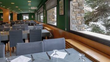 A restaurant with multiple tables and chairs, large windows with a view of snow-covered trees, and stone walls.
