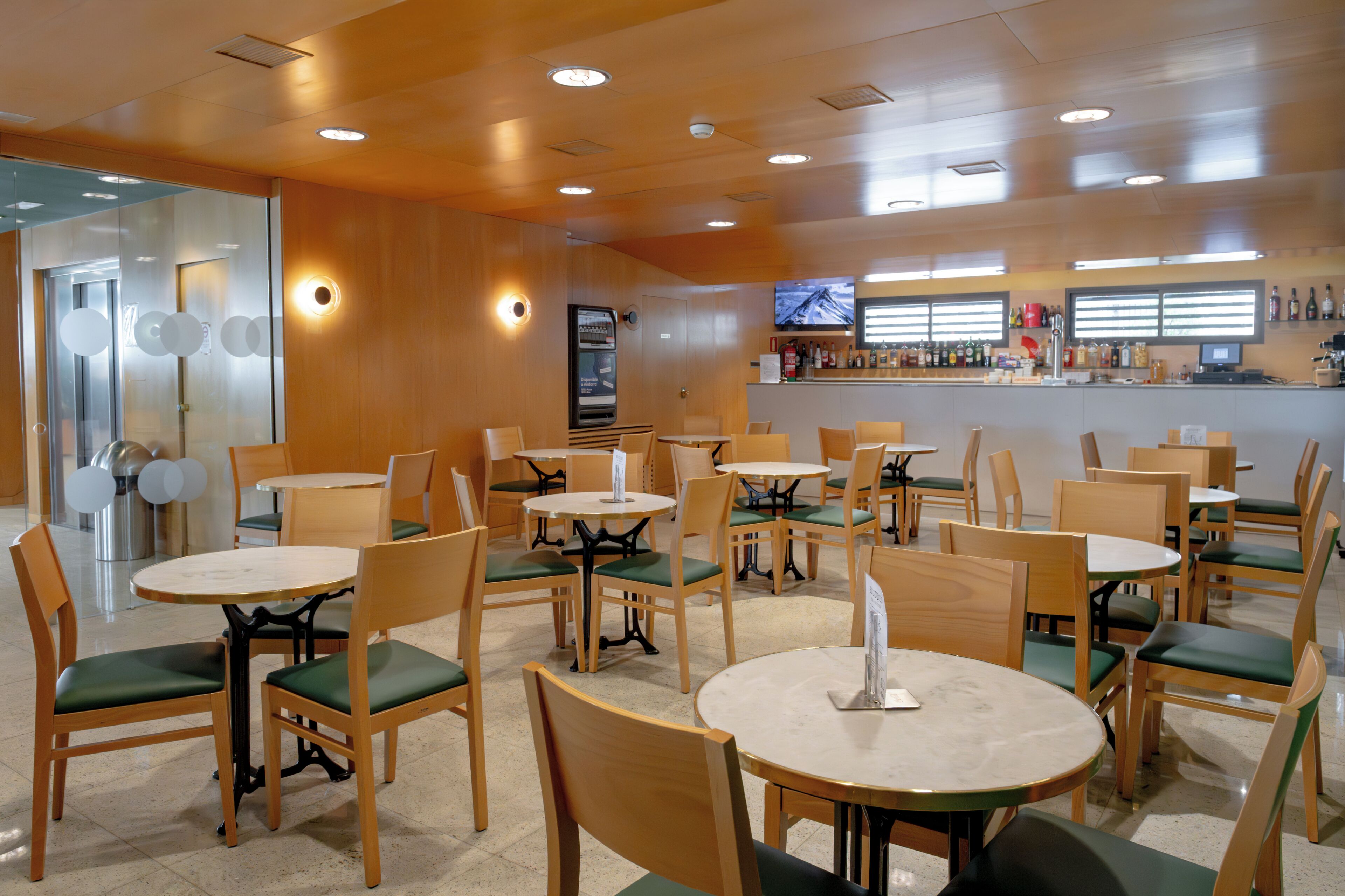 Interior de restaurante con varias mesas y sillas redondas, una zona de bar y una pantalla de televisión en la pared.