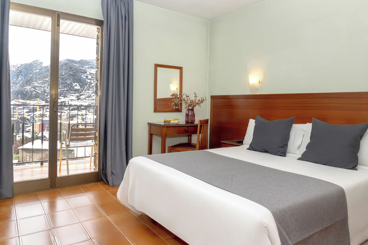 A hotel room with a bed, a desk, a chair, a mirror, and a view of a snowy landscape through a sliding door.