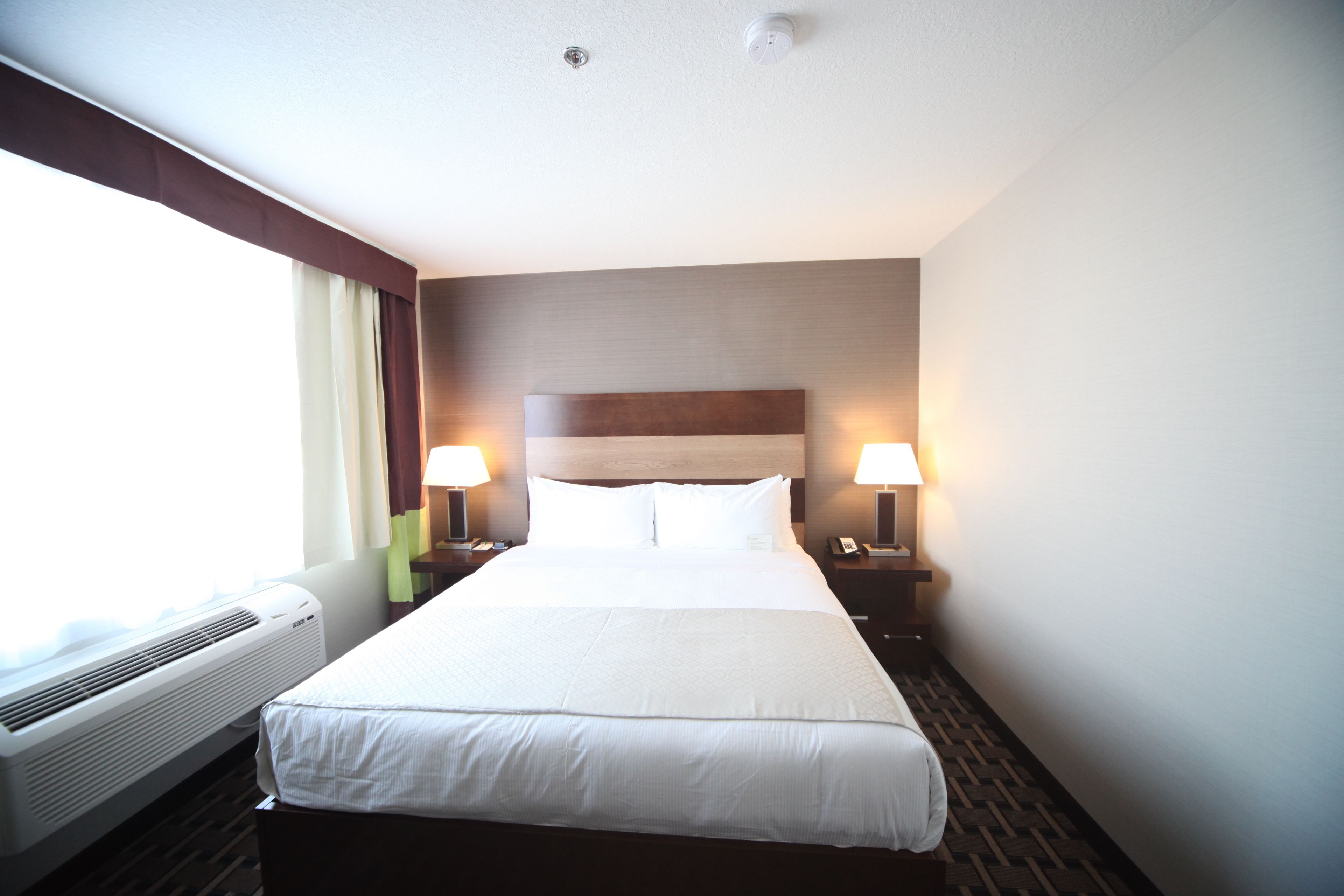 Una habitación de hotel con una cama grande, dos mesitas de noche con lámparas, una ventana con cortinas y aire acondicionado instalado en la pared.
