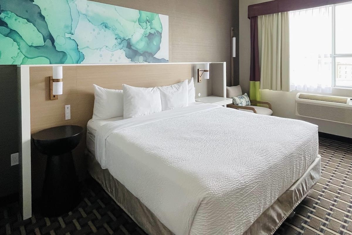 Una habitación de hotel con una cama grande, una mesita de noche, un lavamanos y un baño, y una ventana con cortinas.