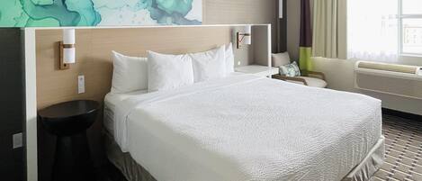A hotel room with a large bed, a bedside table, a nightstand, a wall-mounted lamp, a bathroom sink, and a window with curtains.