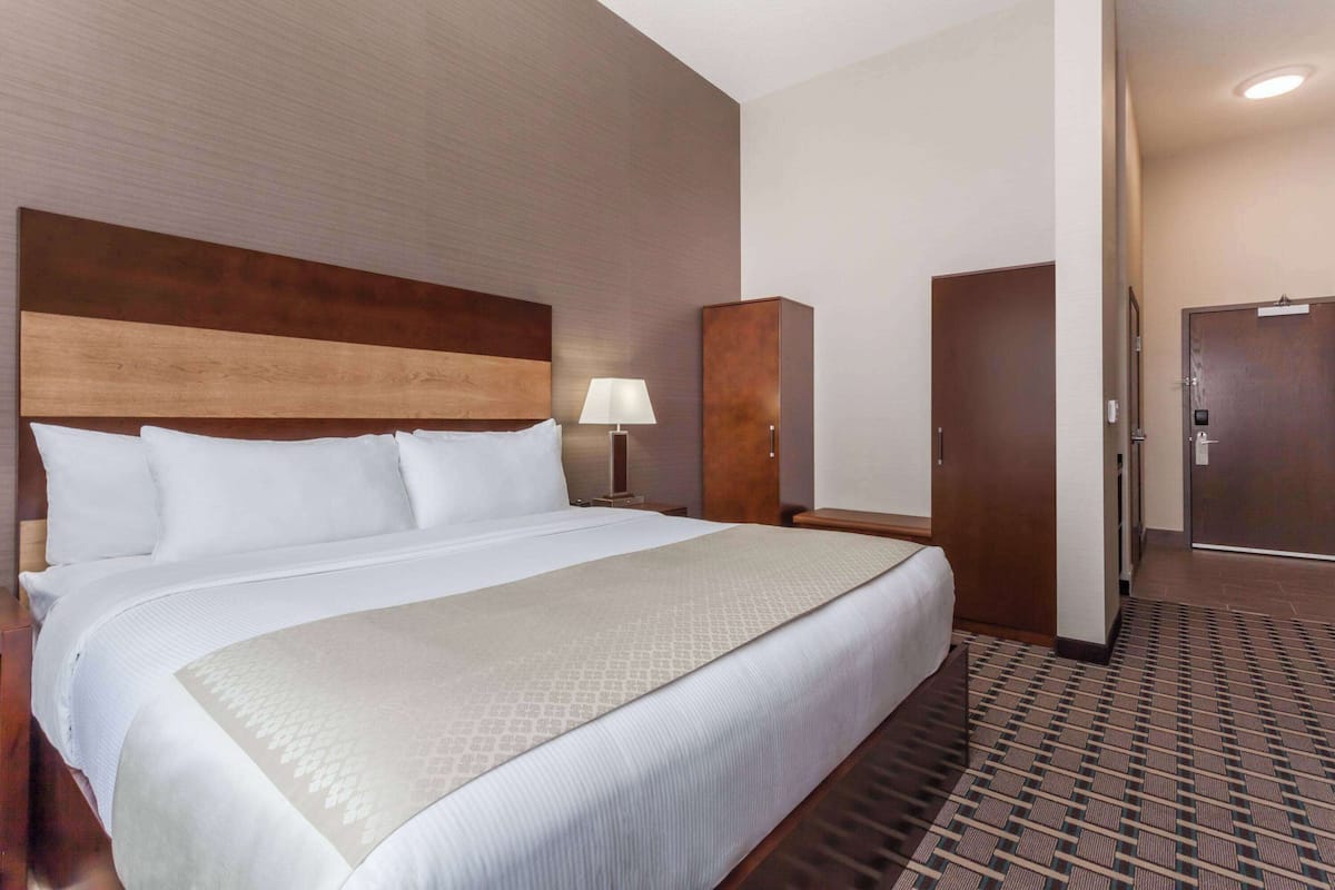 Habitación de hotel con una cama grande, cabecera de madera, mesita de noche con lámpara, armario y una puerta que da a otra habitación.